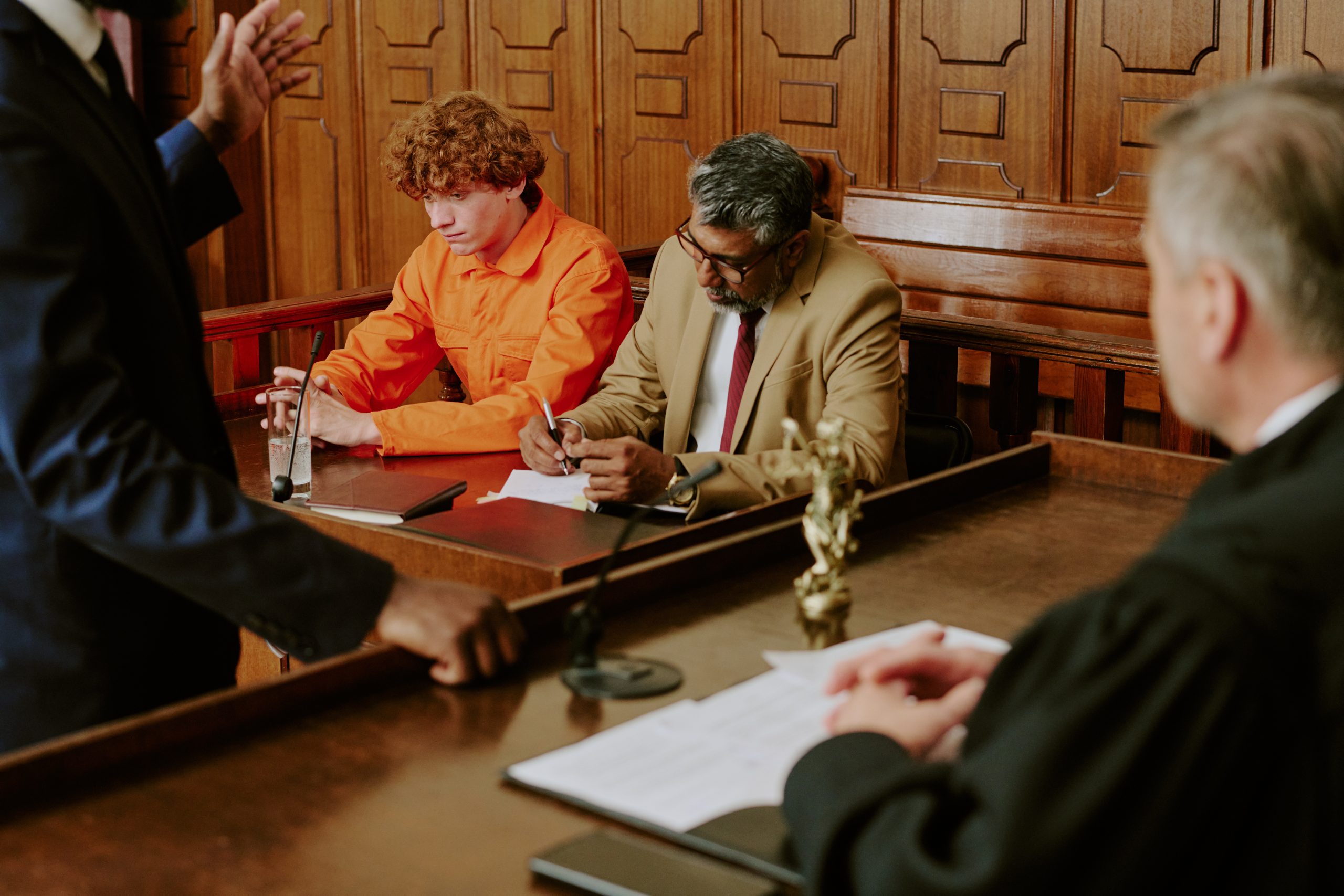 A person in an orange jumpsuit sits beside a lawyer during a courtroom proceeding.
