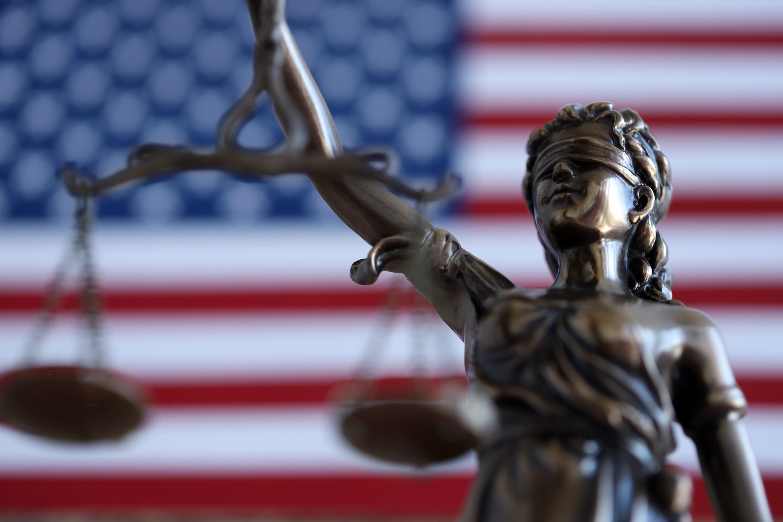A bronze statue of Lady Justice holding scales stands before a blurred American flag background.