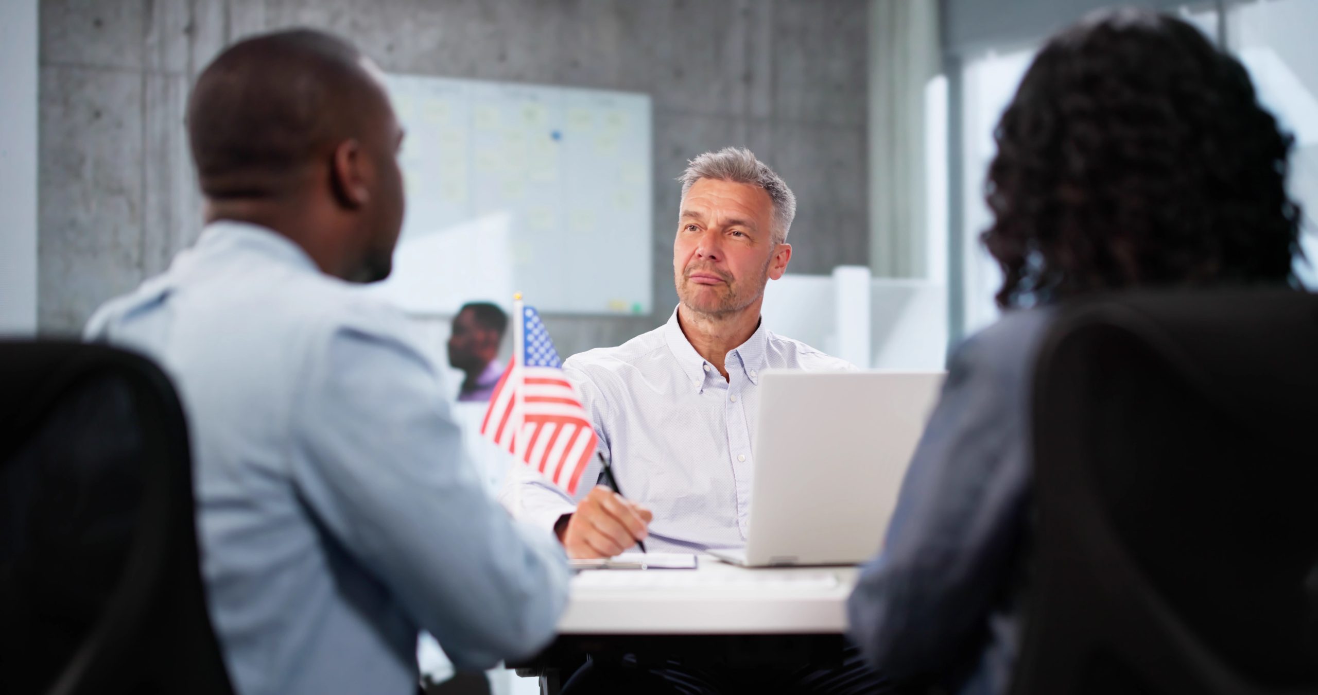 A lawyer sits at a desk with a laptop, attentively listening to two clients during a consultation, with a small United States flag displayed on the table.