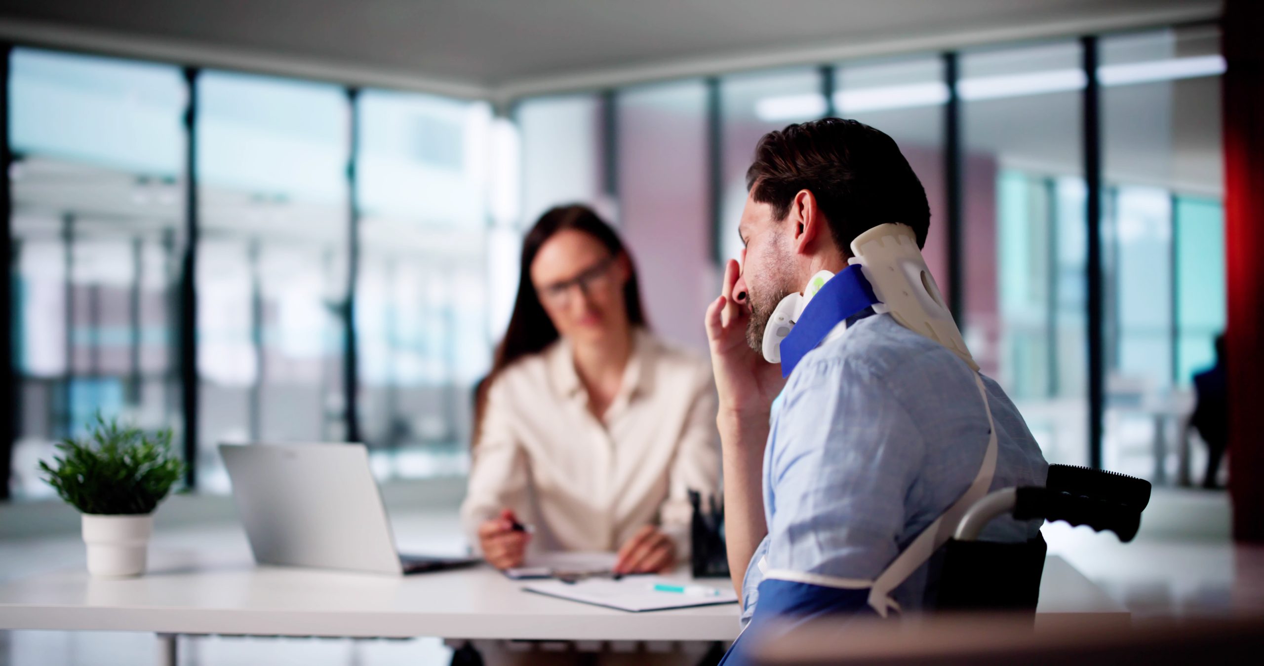 A man wearing a neck brace and arm sling sits at a desk speaking with a lawyer in an office setting, with documents and a laptop on the table.